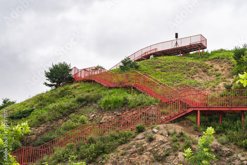  Red metal staircase zigzagging up a greeny hillside unde overcast Sky.