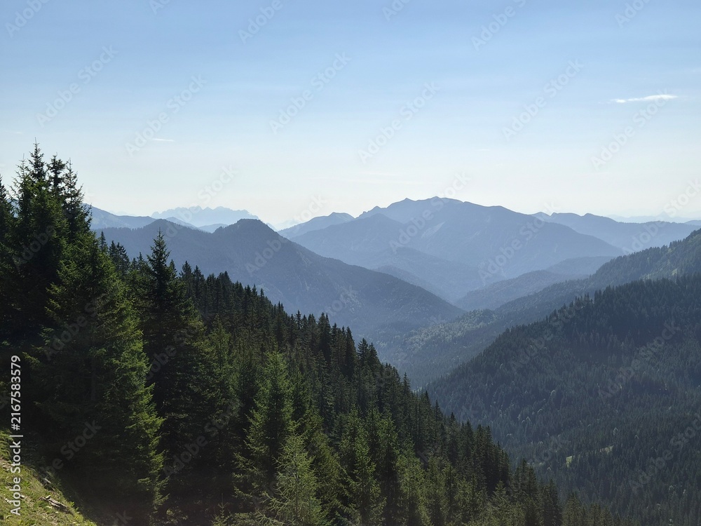 Fototapeta premium Landscape of mountain with a clear sky above a deep forested valley and distant, layered blue Alpine peaks in soft haze. Bird Flying View at Deep Alpine Valley and Mountain Peaks