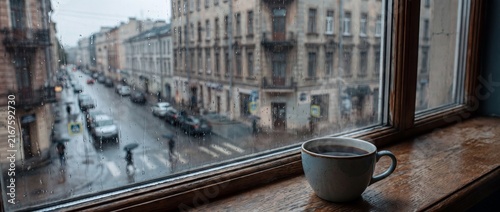 A cozy ceramic cup of hot, steaming coffee resting on a wooden windowsill, looking out at a blurred, moody city street on a rainy day with raindrops on the glass.