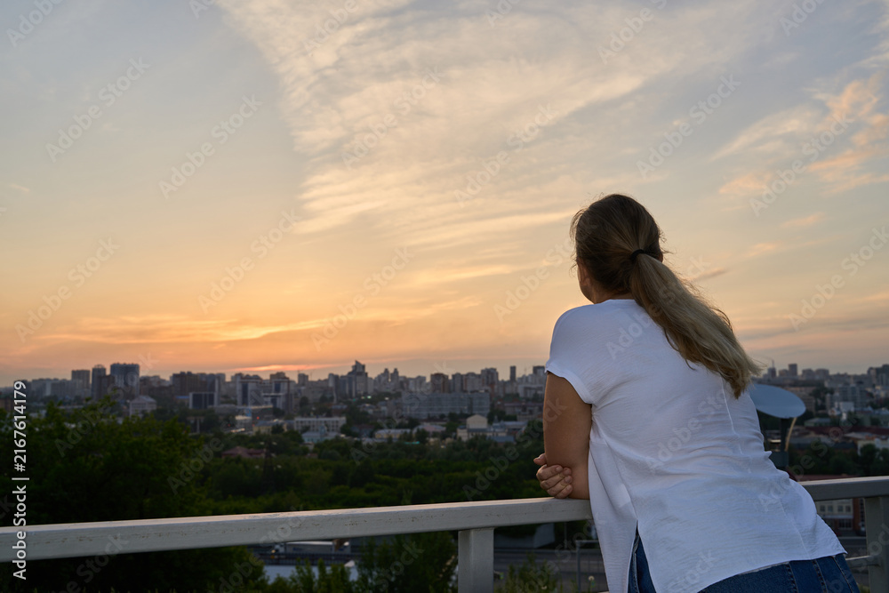Fototapeta premium Young Woman Leaning On Balcony Railing Watching Distant City Skyline Under Warm Sunset Clouds From Elevated Rooftop Terrace.