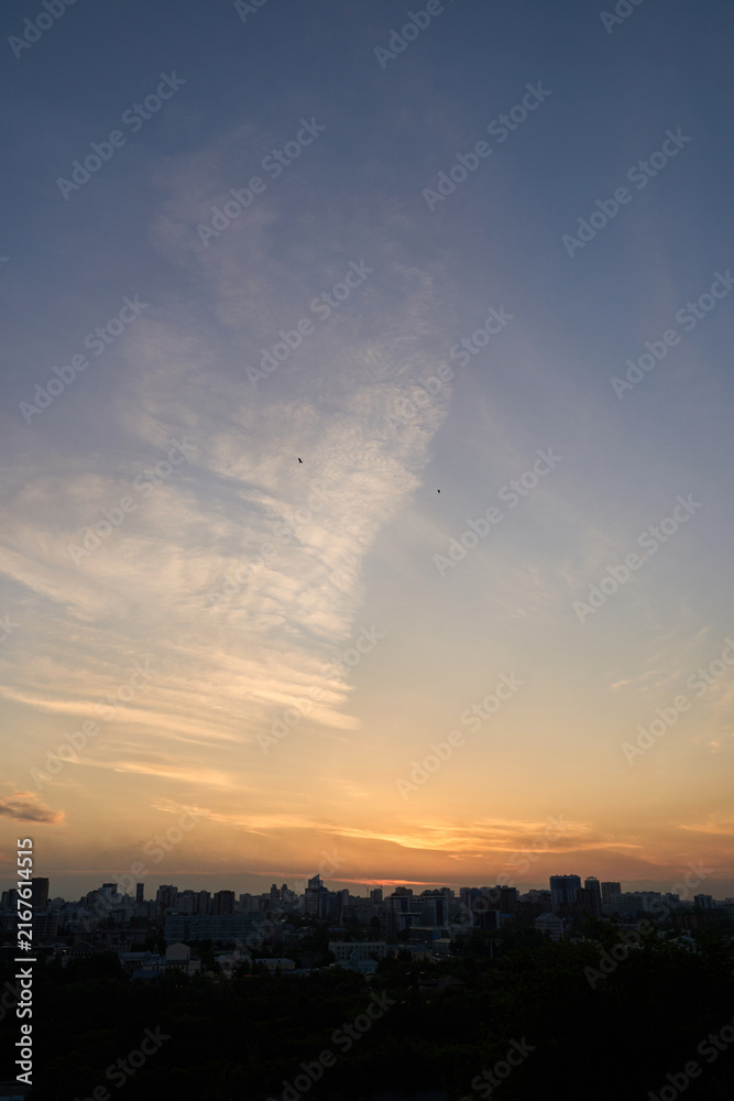 Obraz premium Colorful Sunset Sky Glowing Above Dark City Skyline With Wispy Clouds Over Distant Buildings At Evening.
