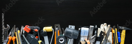 Assorted hand tools, paint brush, gloves, utility knives, pliers, wrench, tape, rulers and hammer arranged in row on black background for repair