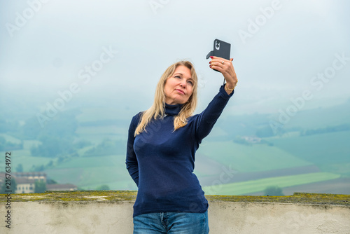 Portrait of a woman standing at an observation point taking a selfie in front of the Langhe Hills on a misty day, Piedmont, Italy