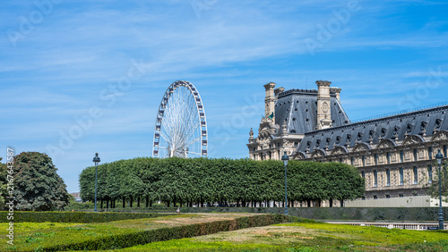 The image shows the garden of Tuileries with a view of the Louvre Museum and the Roue de Paris Ferris wheel in Paris, France
