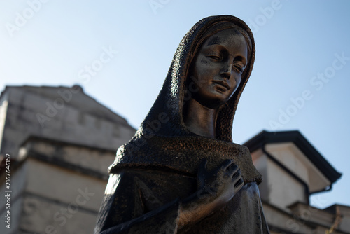 Statua in bronzo della Vergine Maria con mano sul petto in un cimitero monumentale storico, scultura sacra di devozione e fede cristiana su sfondo architettonico