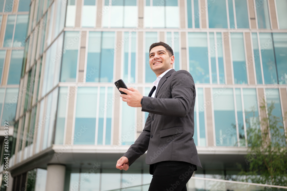 Fototapeta premium Businessman smiles while walking and checking phone outside modern office building