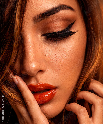 Close up beauty portrait of a sensual female face showcasing flawless glowing skin with freckles, sharp winged eyeliner, and glossy red lips in warm studio lighting.