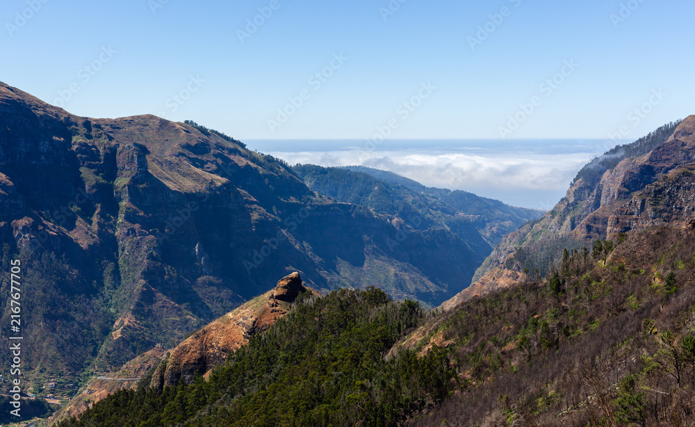 Fototapeta premium Volcanic mountain range dominates Madeira's rugged landscape. Aerial view reveals lush greenery and rocky terrain. Atlantic Ocean meets dramatic cliffs under clear sky.