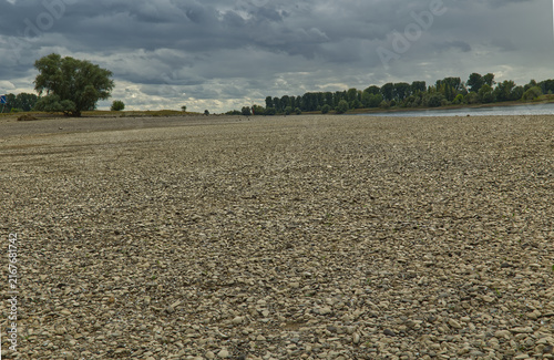 Fotografía Trockenes steiniges Flussufer am Rhein bei Niedrigwasser unter dramatischem Wolk