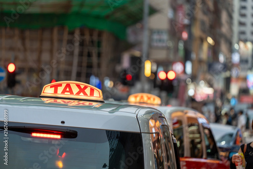 Photography Close-up telephoto view of glowing red taxi rooftop signs on white and red cabs standing in evening city traffic with soft background bokeh