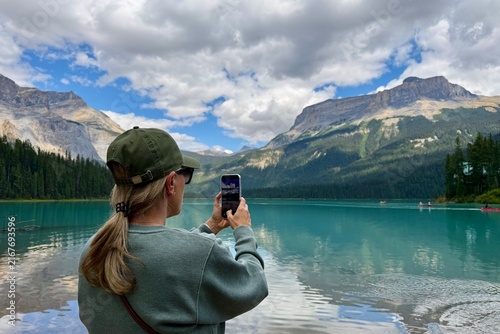 Blonde woman photographed from behind taking photos of turquoise Emerald Lake in Yoho National Park, surrounded by forest and the Canadian Rocky Mountains.