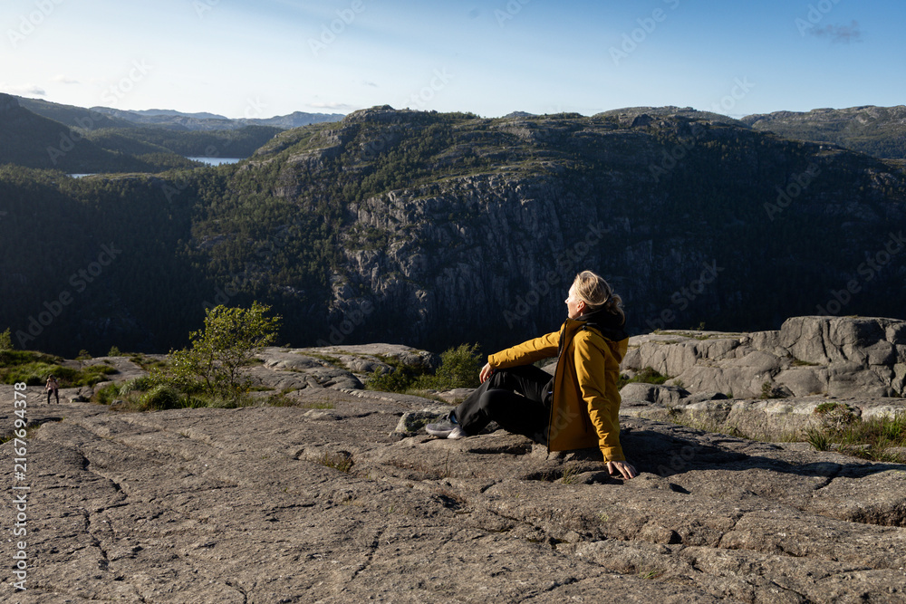 Fototapeta premium Individual wearing a yellow jacket sits on rocky terrain overlooking a vast mountainous landscape with trees and a lake in the distance under a clear blue sky