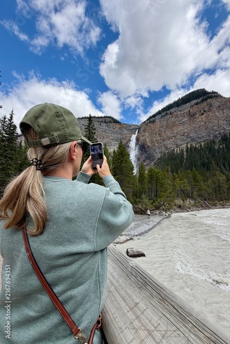Blonde woman photographed from behind taking photos of Takakkaw Falls in Yoho National Park, surrounded by dramatic mountains and forest in the Canadian Rockies.