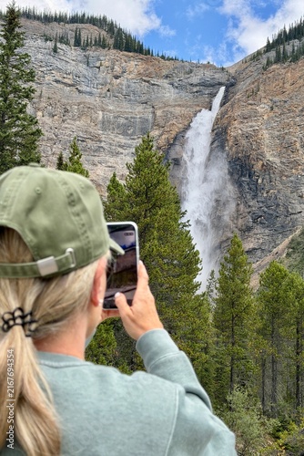 Blonde woman photographed from behind taking photos of Takakkaw Falls in Yoho National Park, surrounded by dramatic mountains and forest in the Canadian Rockies.