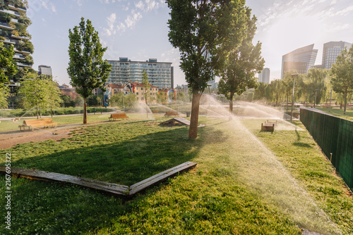 Milan, Italy - May 25, 2026: Green park landscape with trees, benches, and sprinklers watering grass in Milan, modern buildings in background