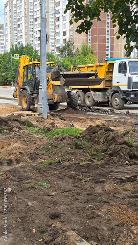 Konstfotografi Yellow backhoe loader digs ground near white dump truck at urban construction site with residential houses