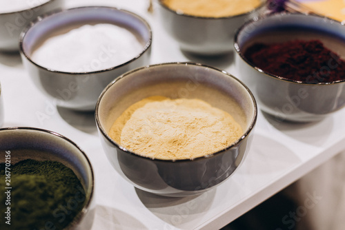 Konstfotografi Assortment of Powders in Bowls: A close-up of several small bowls, each filled with a different colorful powder, arranged neatly on a white surface, hinting at culinary ingredients or craft supplies