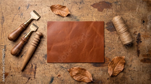 Cognac brown leather piece on rustic wooden workbench with leathercraft tools wooden slicker thread and autumn leaves
