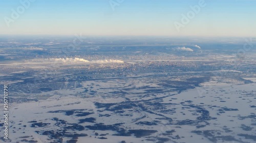 Flying over winter landscape and frozen city of Novosibirsk covered in snow with industrial smoke, aerial view from airplane window, clear blue sky daylight wide shot