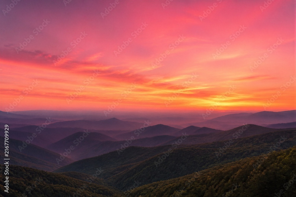 Naklejka premium Sunset paints sky in fiery pink and orange hues. Layered mountain ridges stretch into distant fog. Forested slopes glow softly beneath the glow. Silent majesty evokes peace and awe
