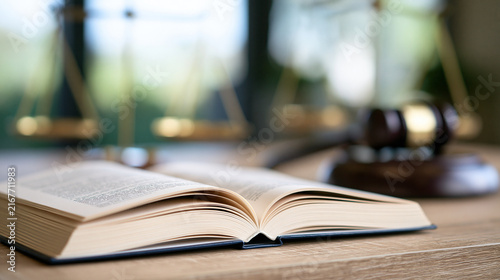 An open law book rests on a wooden desk with a blurred gavel and scales in the background. Use this legal concept image for law firm websites and courtroom editorials.