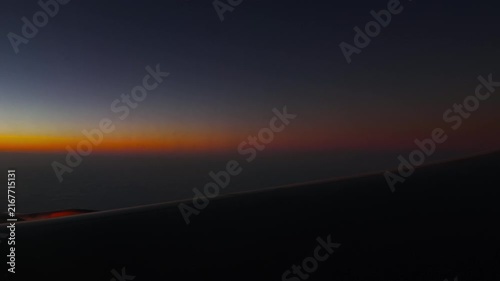 Airplane wing flying above cloud layer during colorful sunset at high altitude, scenic view from passenger window at dusk with orange sky gradient