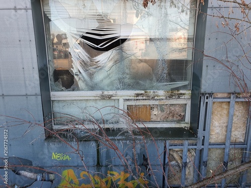 A photograph depicting a severely damaged window on an urban building facade. The window glass is extensively cracked and shattered with a large central hole, revealing black and white striped