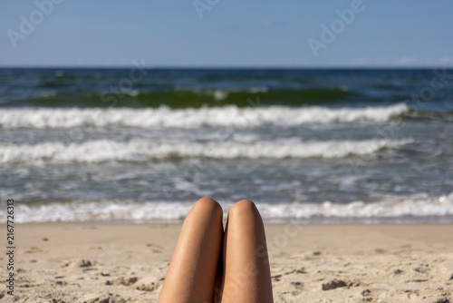 Woman's Tanned Legs Relaxing on Sandy Beach Facing Ocean Waves