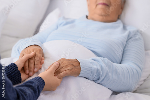 Caregiver with lying senior woman holding hands in bedroom, closeup. Care con...