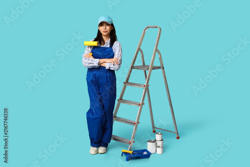Female Asian painter with rollers, paint cans, tray and stepladder on blue ba...