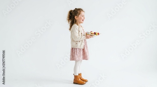 Joyful young girl in cream cardigan and pink floral dress, profile view, holding plate of colorful sweets, smiling happily against white studio background