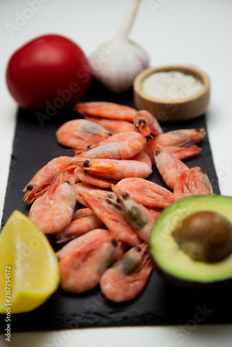 Frozen shrimps, a slice of lemon, half an avocado are laid out on the black slate, a tomato, garlic and a salt shaker are blurred in the background. Concept: still life with seafood and fresh