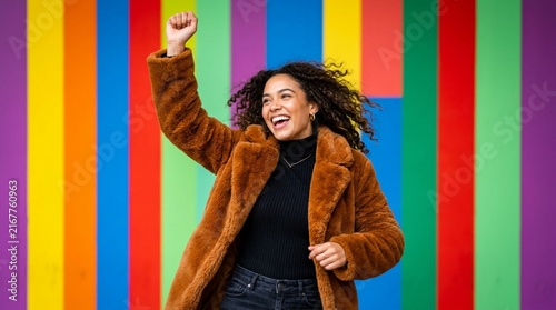 Joyful African American woman in cozy brown faux fur coat raising fist in celebration against vibrant multicolor striped backdrop, expressing happiness and triumph in bold colorful lifestyle portrait