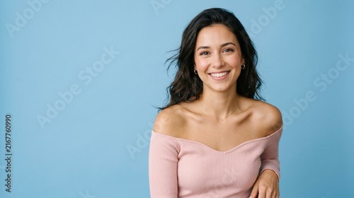 Smiling young brunette woman in soft pink off-shoulder top posing against clean sky blue studio background with natural makeup and wavy hair