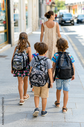 A happy mother with three little kids walking down a city street on a sunny day, going back to school, traveling together or heading to an education class with backpacks