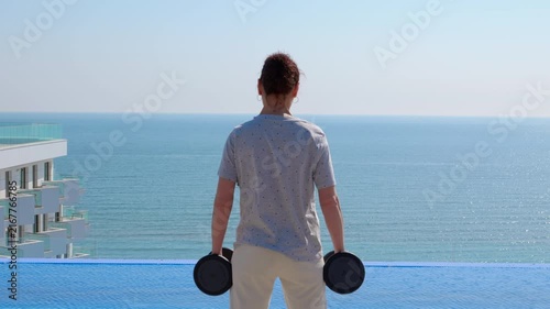 Woman training with dumbbells overlooking the ocean. Athletic woman performing bicep curls with dumbbells while standing near an infinity pool, enjoying a strength training workout