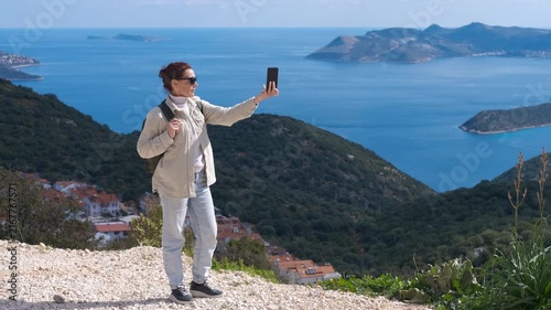 Female traveler taking a selfie on a mountain top overlooking the sea. Young female traveler taking a selfie with her smartphone from a mountain viewpoint, capturing the stunning coastal landscape