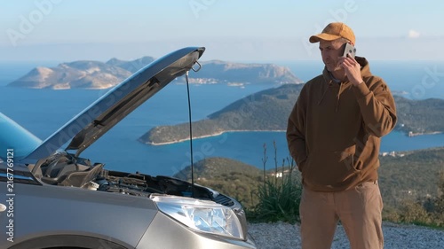 Stressed man calling roadside assistance for broken car. Worried man standing by his broken down car with an open hood on the side of a scenic mountain road, anxiously talking on his smartphone