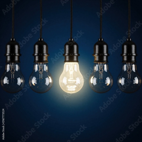One light bulb glows brightly among unlit ones on a dark background. Concept represents unique idea, innovation, or standout solution in a group.