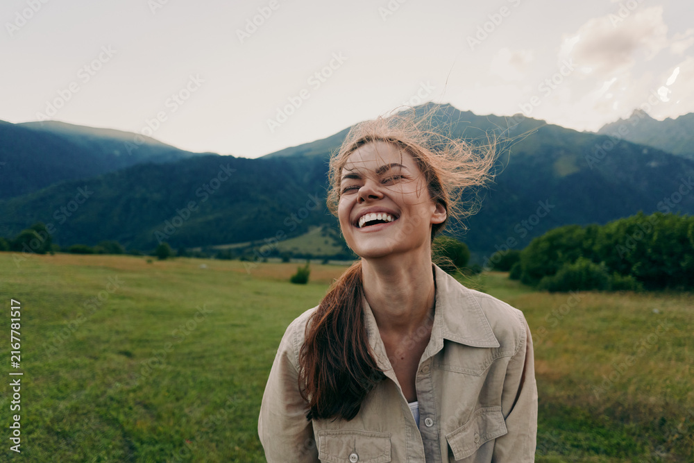 Fototapeta premium A smiling young woman stands in a green meadow with mountains in the background, wind in her hair, capturing a carefree outdoor moment, with light suggesting freedom today.