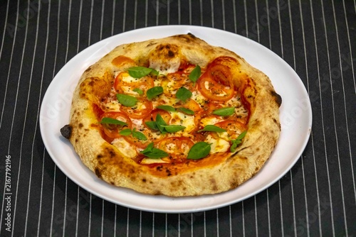Neapolitan-style personal pizza with naturally leavened long-fermentation dough, sliced tomatoes, melted mozzarella, fresh basil and oregano, served on a white plate over a dark pinstriped cloth.