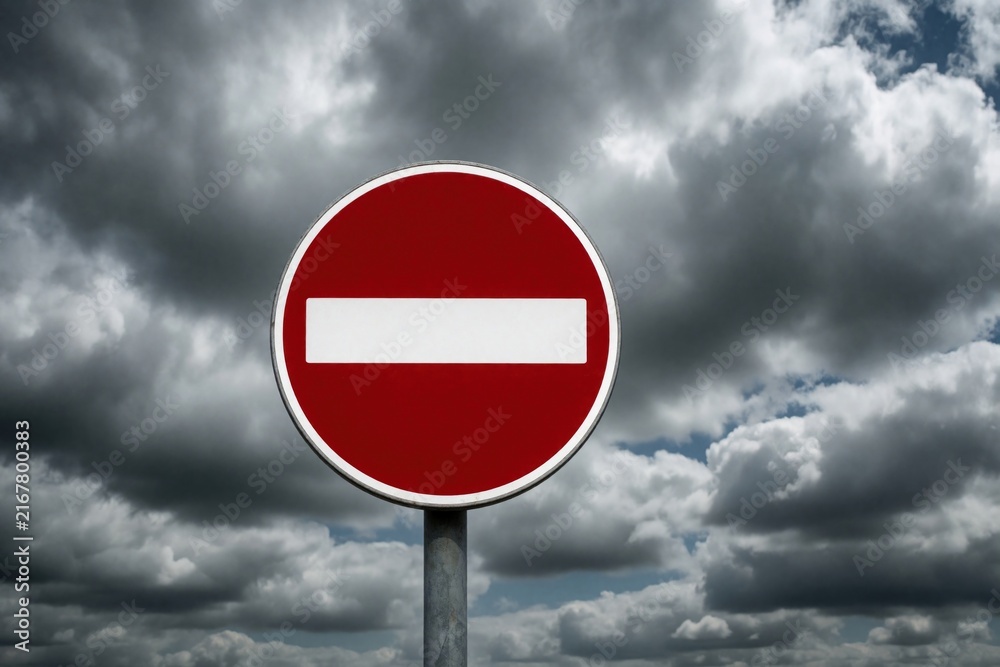 Fototapeta premium Red no entry road sign stands against dramatic cloudy sky. Round traffic symbol indicates forbidden direction street restriction. Warning sign shows stop signal for drivers under dark stormy clouds.