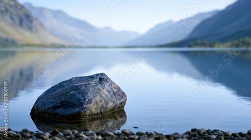 A large rock immersed in calm reflective lake water with serene mountain ranges softly blurred in the distant background under clear sky