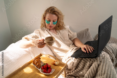 A blonde woman working in bed with green eye patches, typing on her laptop while holding a cup of coffee next to a breakfast tray with croissants and strawberries
