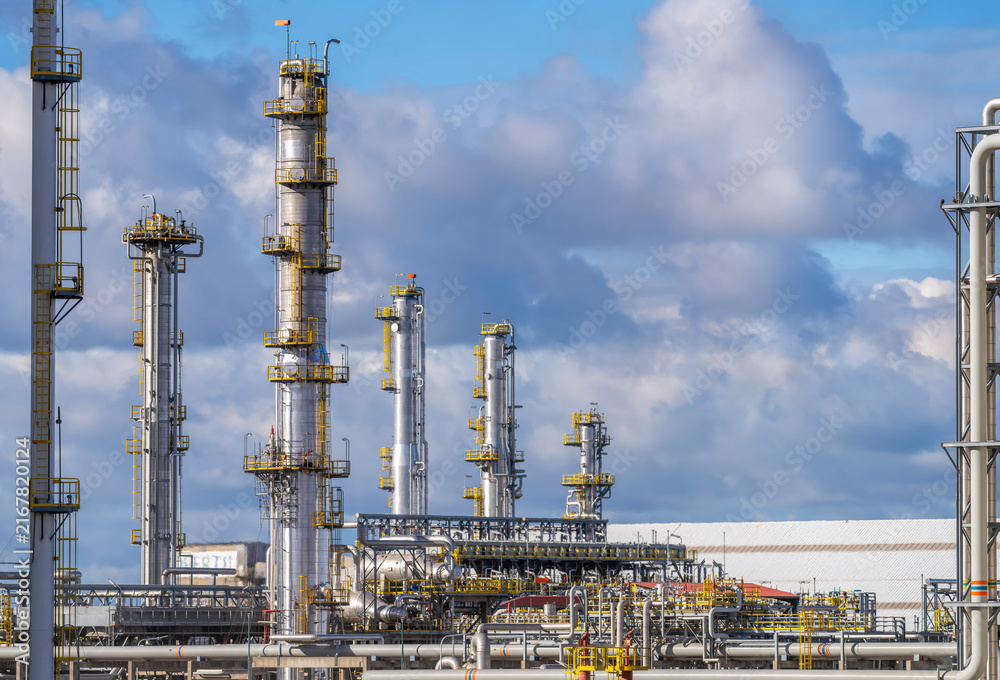 Obraz premium Complex petrochemical refinery plant with tall steel distillation columns, metal pipes, intricate industrial structures under bright blue sky with fluffy white clouds