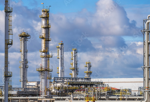 Complex petrochemical refinery plant with tall steel distillation columns, metal pipes, intricate industrial structures under bright blue sky with fluffy white clouds