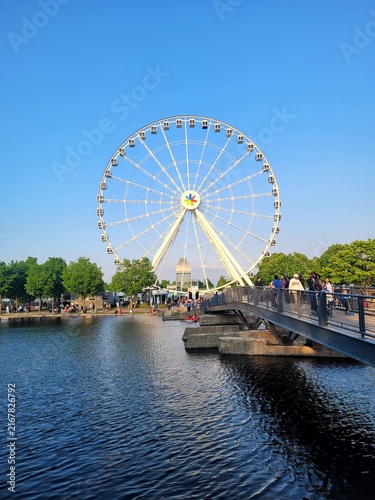 Large observation Ferris wheel standing behind a footbridge and pond in a city park on a clear sunny day under a blue sky.