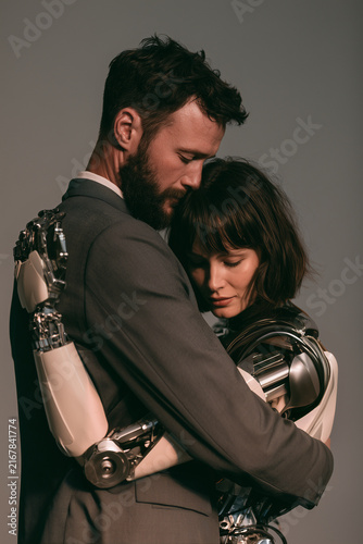 man hugging female cyborg ai robot companion isolated on plain gray studio background