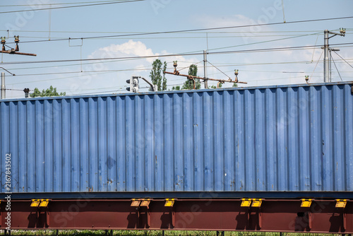 Blue shipping container carried on a cargo train beneath railway power lines, showing intermodal freight transport, rail logistics infrastructure and containerized goods movement across the network.