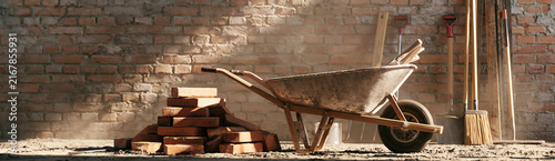 A rustic wheelbarrow rests on a concrete floor beside a stack of red bricks and construction tools against a rough brick wall.